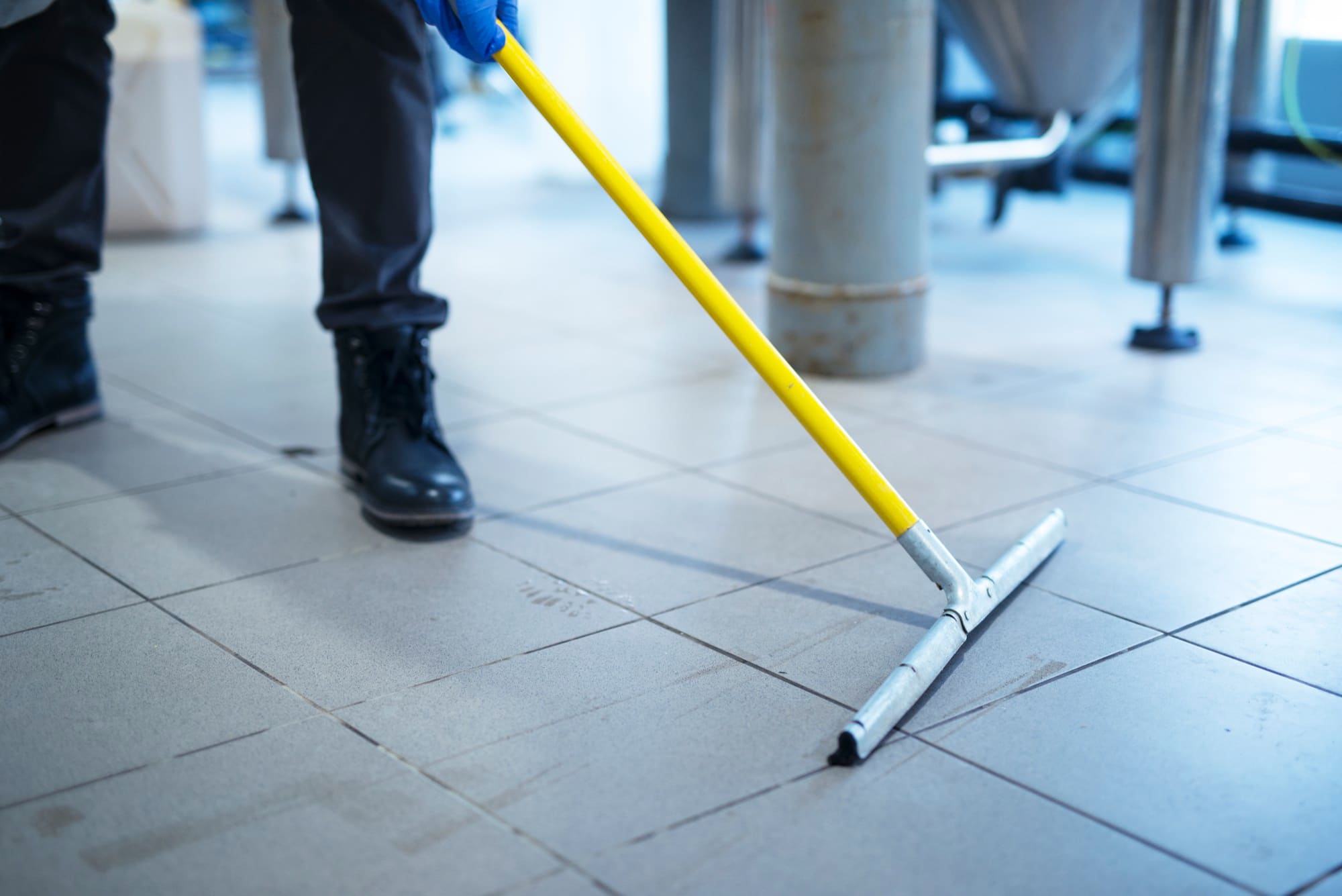 A person wearing black boots and gloves uses a yellow-handled floor squeegee to clean a tiled floor in an industrial or commercial setting, demonstrating the thorough attention to detail provided by professional Construction Cleaning Services.