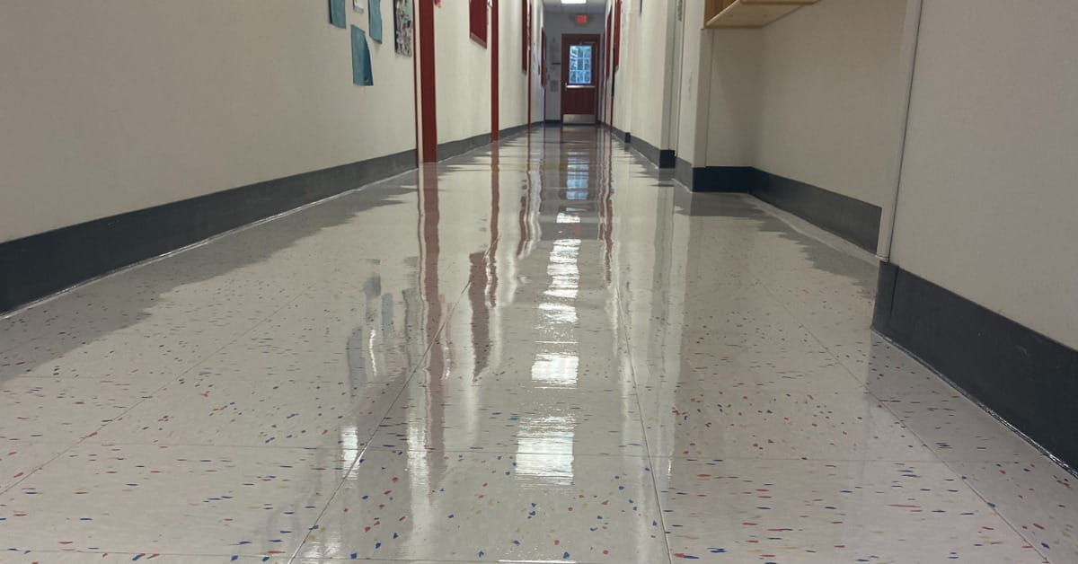 A shiny, freshly waxed hallway floor with white tiles and colorful specks, white walls, and red door frames. Art is pinned to the wall on the left—an impressive example of Commercial Cleaning that highlights the high standards maintained in Westland MI.