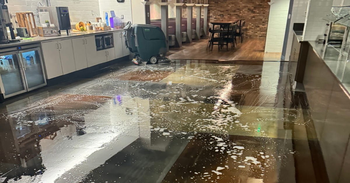 The interior of a cafe or food service area during cleaning, with a green floor scrubber on a wet, soapy floor. The foreground features reflections of lights and counters, while the background shows seating areas, counters, and brick walls in a well-lit space.