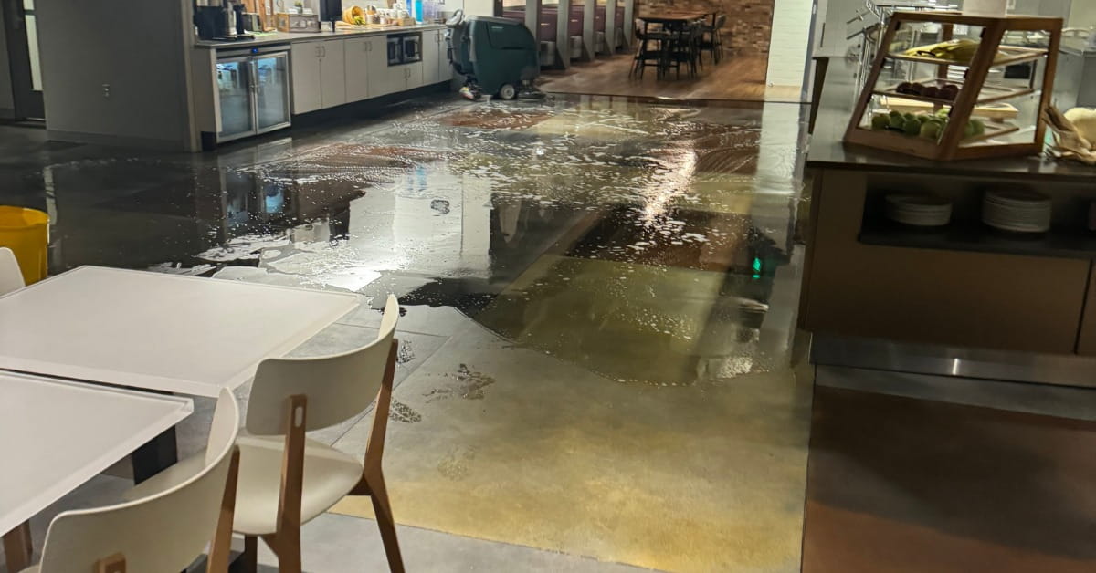 A cafe or dining area with a wet, soapy floor reflecting overhead lights and furniture. A green floor cleaning machine is visible near the center, and wet footprints lead across the floor. The background includes counters with food displays, tables, and chairs, suggesting an ongoing cleaning process.