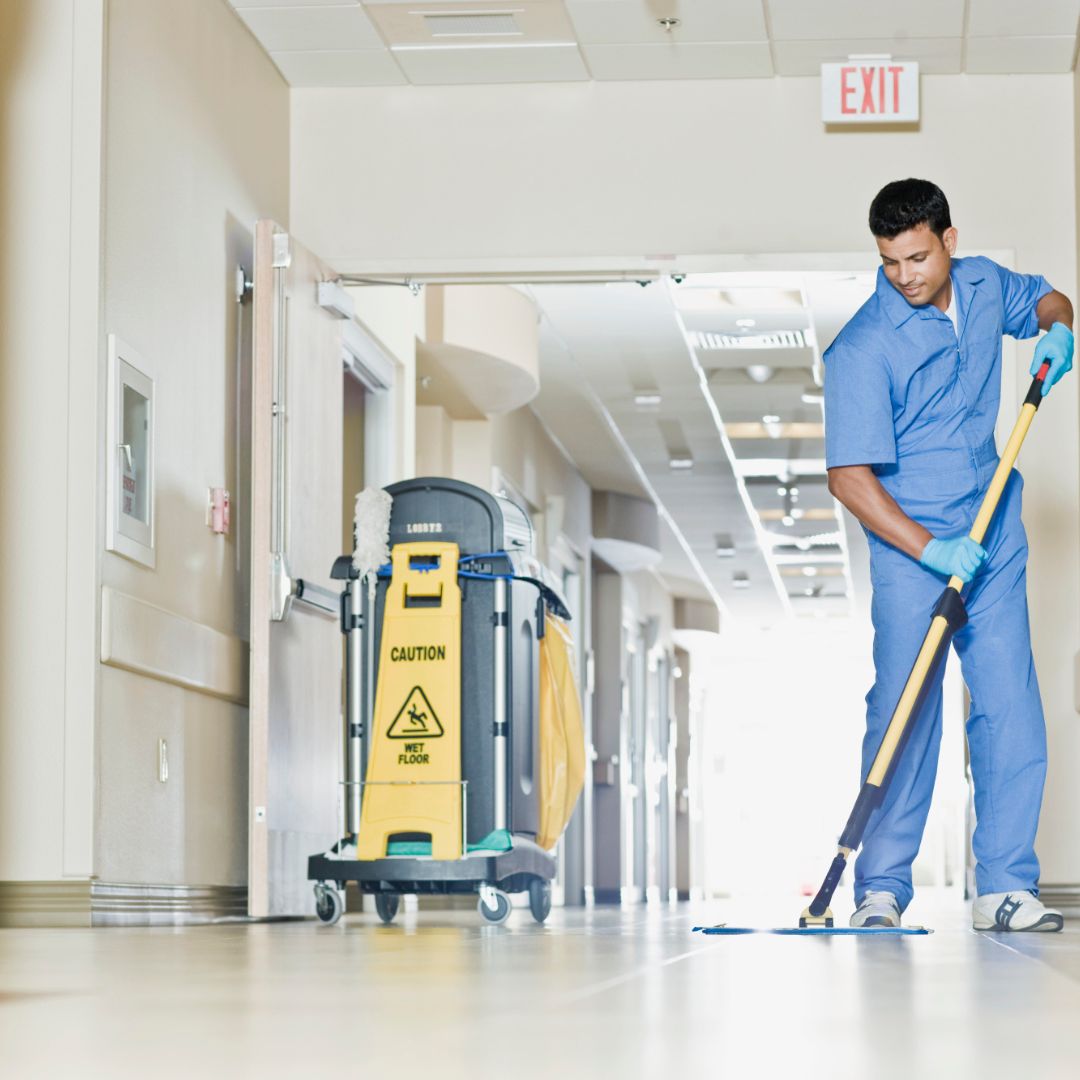 A male custodian in a blue uniform and gloves mops a light-colored, reflective hallway floor. A cleaning cart with a yellow 