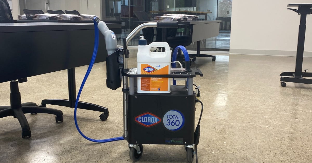 A Clorox Total 360 disinfectant sprayer system on a black cart with a white Clorox Disinfectant Cleaner bottle on the top shelf. The cart features a sprayer unit with blue hoses and is positioned in a modern office or conference room with a black table, chairs, and light-colored flooring.