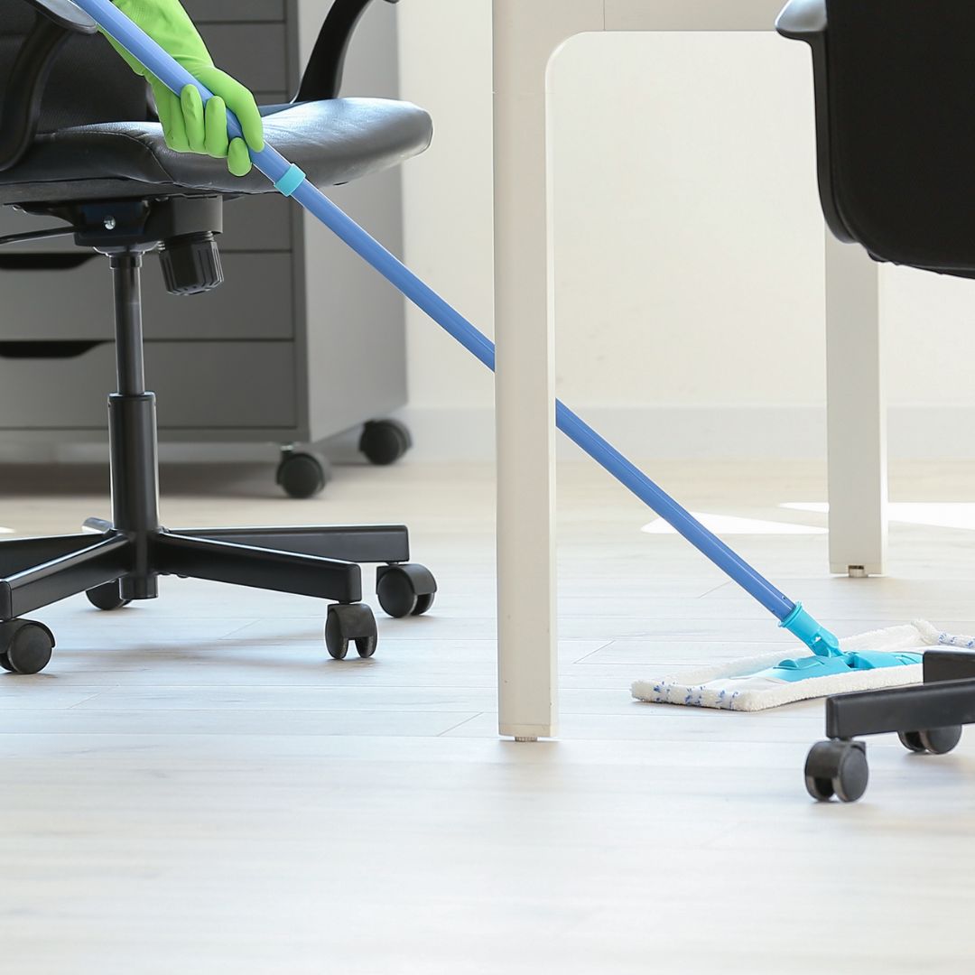 A person wearing a green glove is mopping a light-colored wooden floor in an office setting. The blue-handled mop and black office chairs are prominent, with a white desk leg and a filing cabinet in the background.