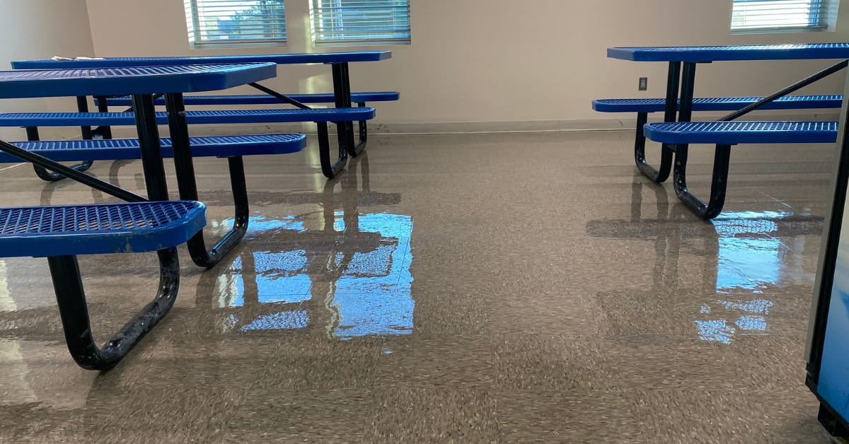 An empty indoor cafeteria or common area with blue picnic-style tables and benches on a glossy, light brown tiled floor. The floor reflects the tables and overhead lighting, while windows with blinds allow natural light into the clean, well-maintained space.
