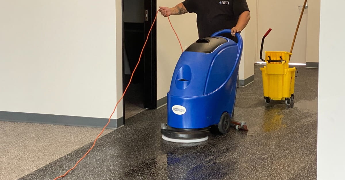 A person wearing a black shirt with 'BRETT' on the back operates a blue floor scrubber in a commercial hallway. A bright orange power cord trails behind the machine, and a yellow mop bucket with a wringer is visible nearby. The floor is partially wet and reflective, contrasting with the dry sections ahead.