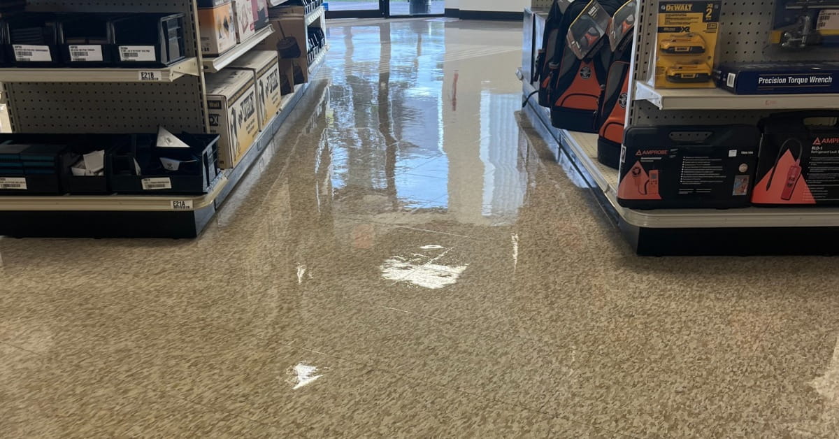 An aisle in a hardware store with metal shelving units on both sides, displaying tools and components. The light brown tiled floor is highly reflective, mirroring the overhead lights and distant entrance, creating a bright and orderly commercial environment.