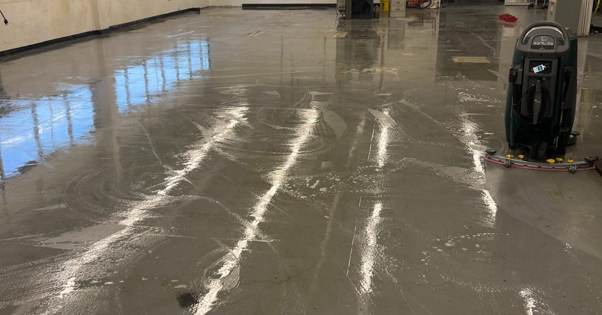 A wet, reflective gray floor in a commercial or industrial space being cleaned by a teal floor cleaning machine. The floor shows streaks and foam patterns from the cleaning process, with prominent reflections of windows and bright lighting. The cleaning machine features a red-edged squeegee bar and a control panel, positioned on the right side of the frame.