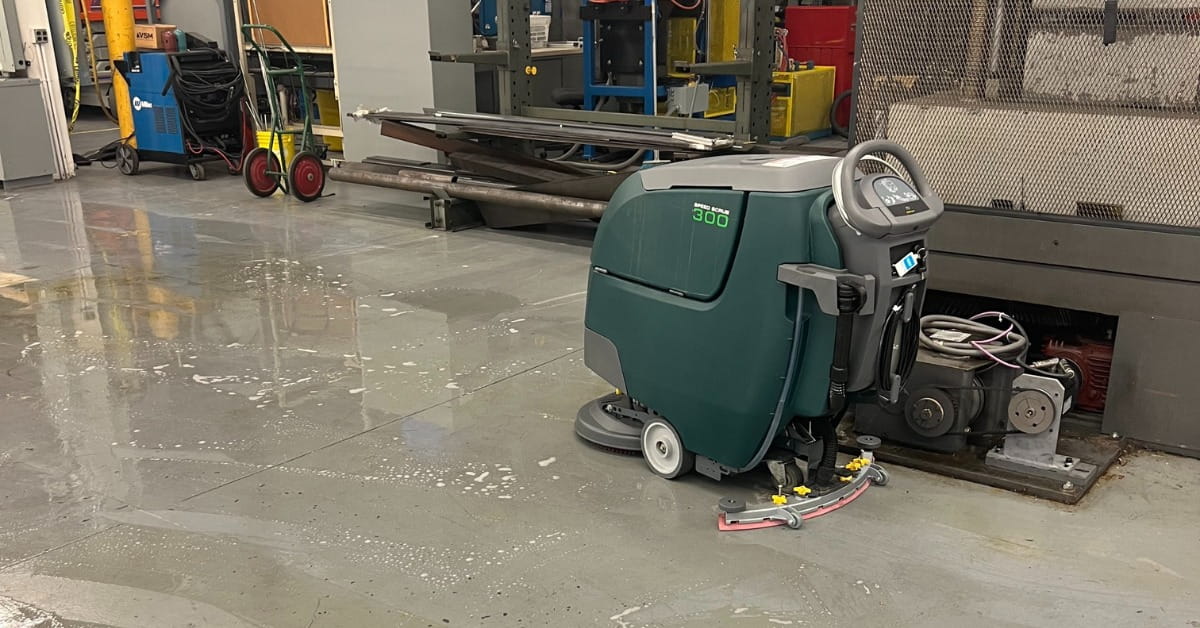 An industrial workshop with a green floor cleaning machine in the foreground, actively cleaning a gray concrete floor. The floor is wet with soapy residue, and the background features a blue welding machine, metal storage racks, and other industrial equipment.