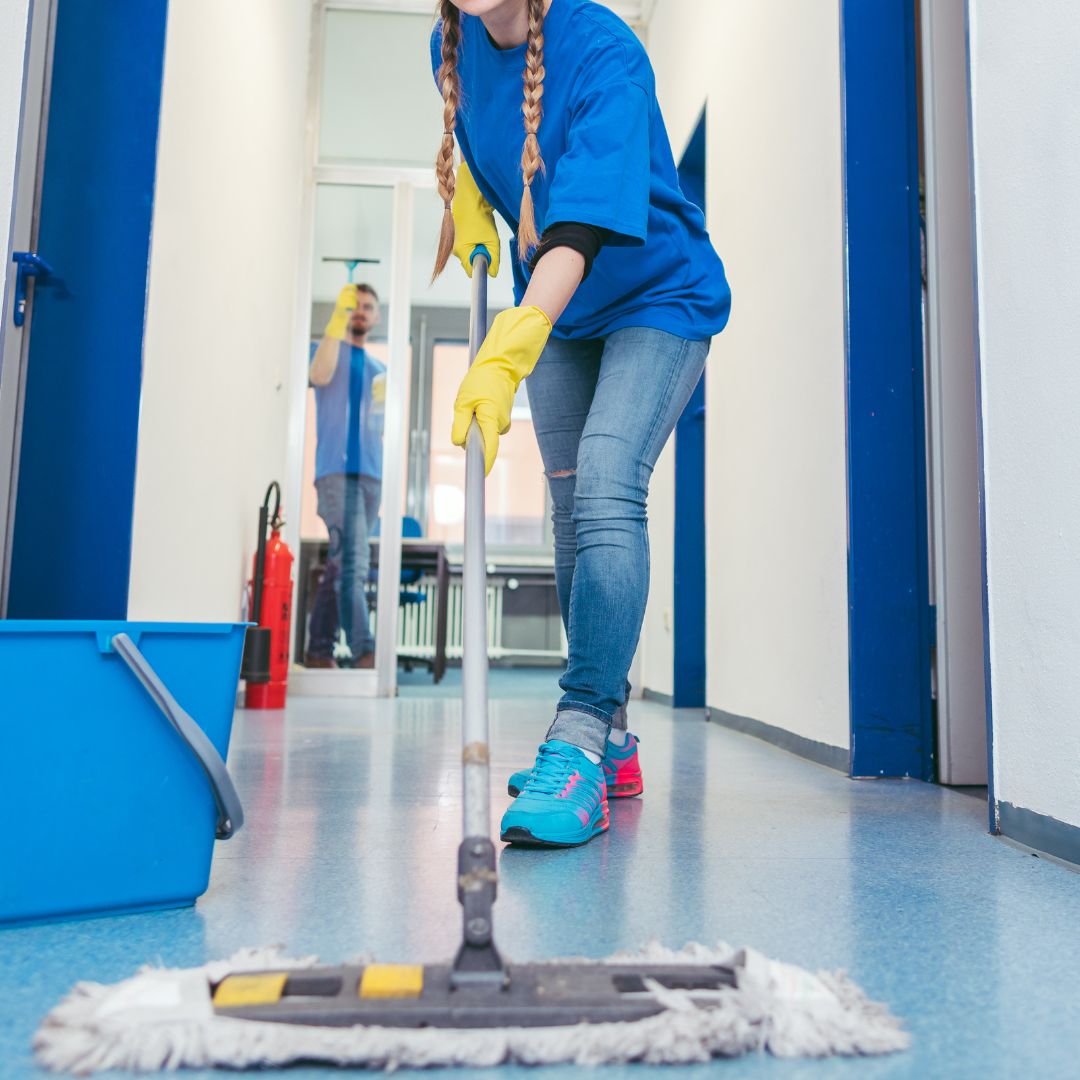 Two people cleaning a brightly lit corridor. A woman in the foreground is mopping the blue-tiled floor, wearing a blue shirt, ripped jeans, and yellow gloves. A man in the background is cleaning a window with a squeegee.