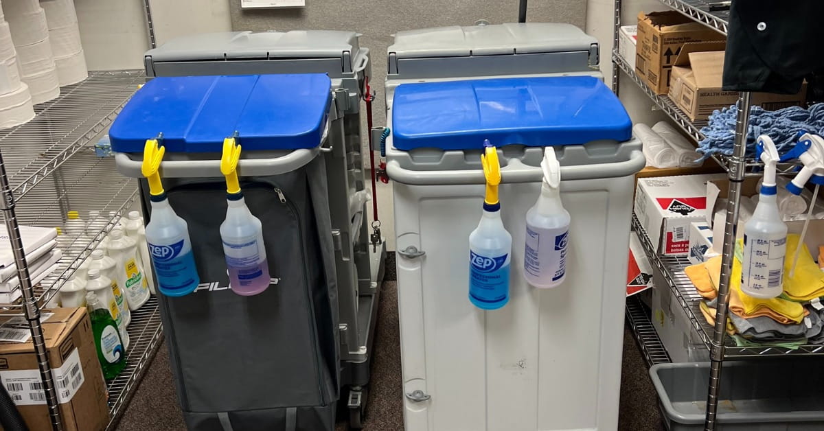 A well-organized janitorial supply room featuring two professional cleaning carts with spray bottles clipped to their sides. Surrounding metal wire shelves are stocked with cleaning supplies, including Zep Professional products, paper towels, mop heads, and cardboard boxes. The floor is carpeted, and the space is brightly lit.