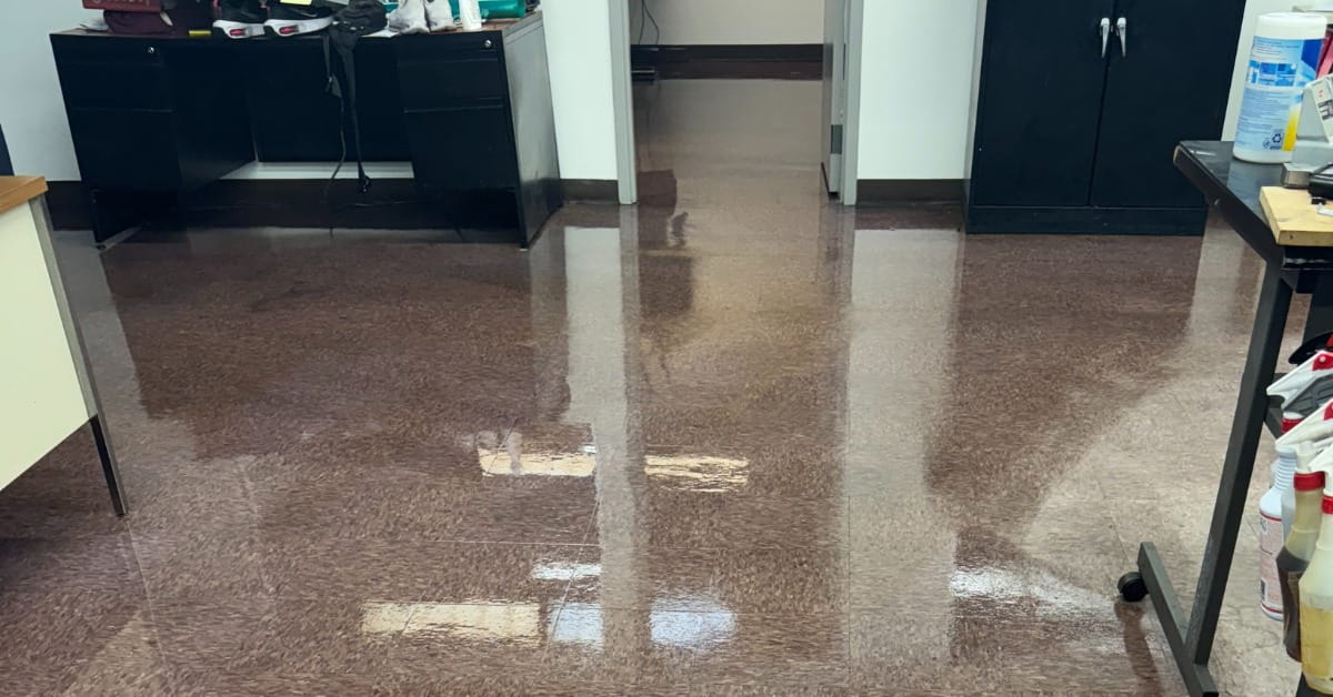 A polished, reflective floor in a commercial or office space with a speckled terrazzo-like pattern. Reflections of overhead lights, furniture, and doorways are visible. The room includes dark cabinets, a desk with personal items, and a table with bottles and cleaning supplies, suggesting a functional workspace.