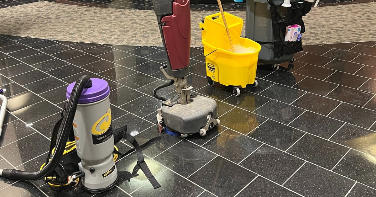A collection of professional cleaning equipment, including a gray and purple backpack vacuum, a red and gray floor buffer, a yellow mop bucket with a wringer, and a gray cleaning cart. The equipment is positioned on a shiny black tiled floor next to a light gray carpeted area, indicating active or recent cleaning in a commercial space.