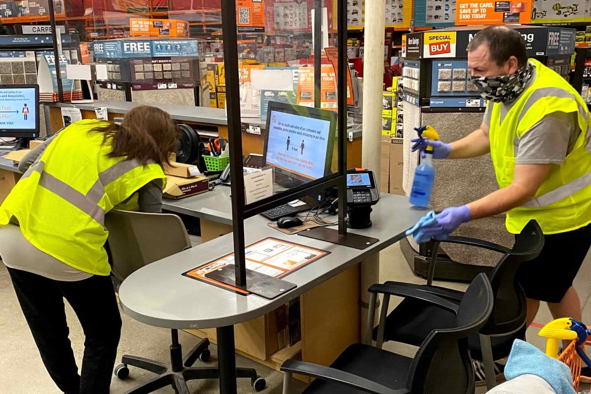 Two employees in yellow vests clean and disinfect a checkout area at a store, ensuring a safe and welcoming environment for everyone. One sprays cleaner while the other wipes surfaces with care. A computer monitor displays a social distancing graphic, highlighting new safety protocols prominently featured on the homepage.