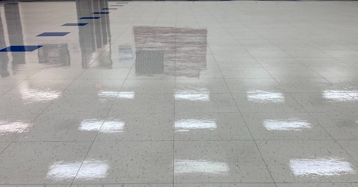 A dark, highly reflective floor resembling polished concrete or tile, mirroring a doorway or hallway with light-colored walls and a baseboard. The reflections include vertical lines and subtle marks, emphasizing the floor's glossy surface and the room's clean, minimalistic design.