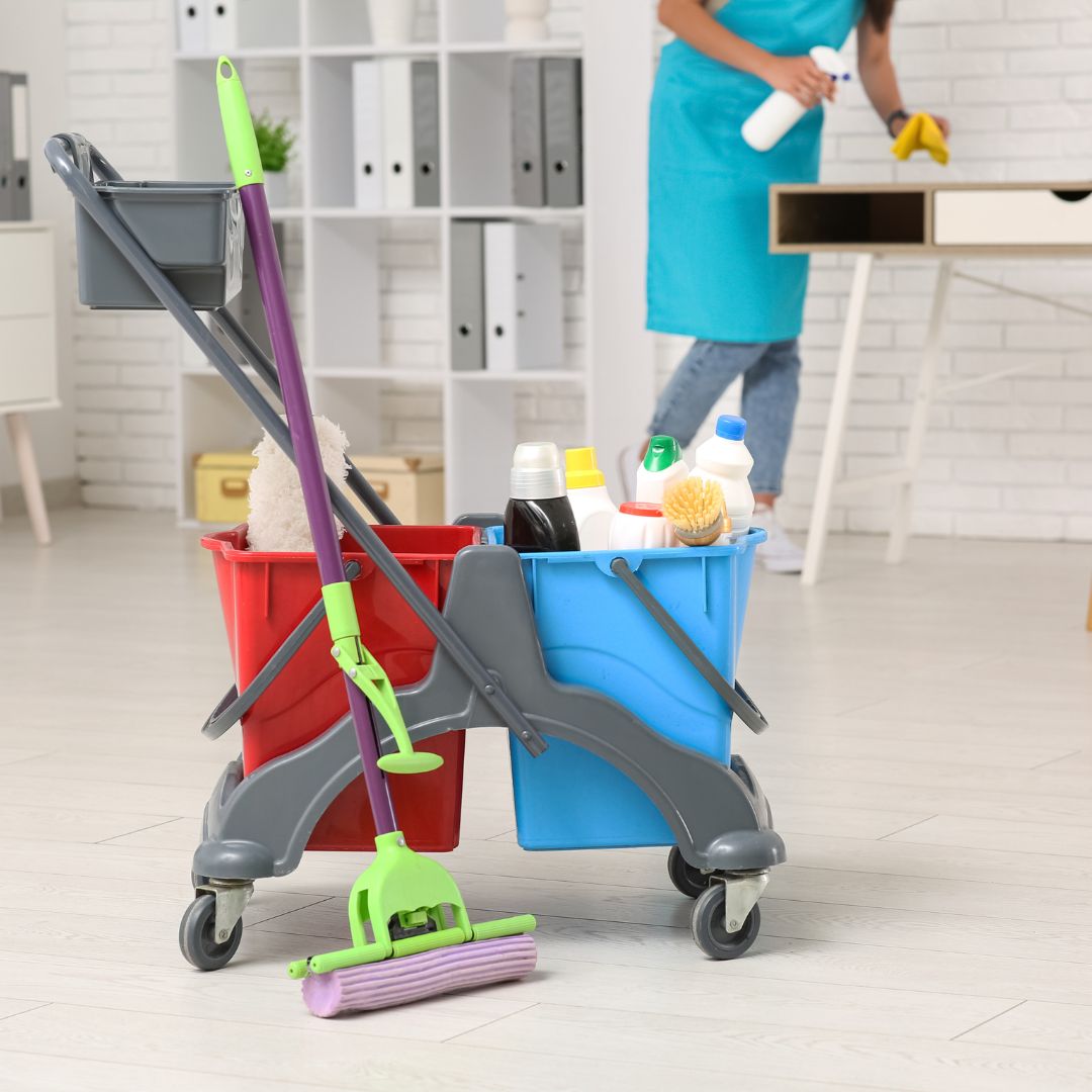 A cleaning cart with red and blue buckets, a mop, and various cleaning supplies is in focus in an office setting. In the blurred background, a person wearing a teal apron cleans a desk with a spray bottle and a yellow cloth. The scene emphasizes cleanliness and maintenance.
