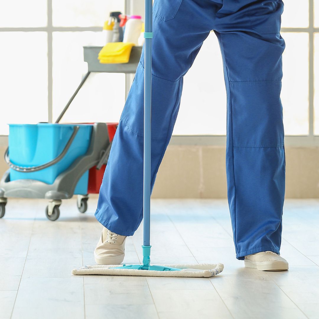 A close-up of a person in blue work trousers and light-colored shoes mopping a light wood or tiled floor. A professional cleaning cart with blue and red buckets and cleaning supplies is visible in the background, set against a bright window.