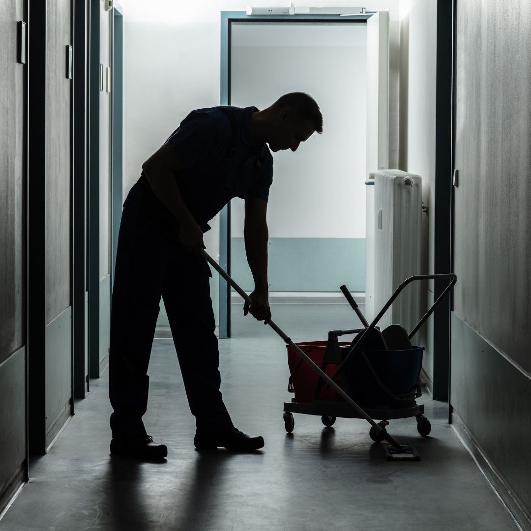 A silhouetted cleaning worker mopping a reflective hallway floor. A cleaning cart with red and blue buckets is nearby, and the scene is backlit by light streaming from an open door at the end of the hallway.