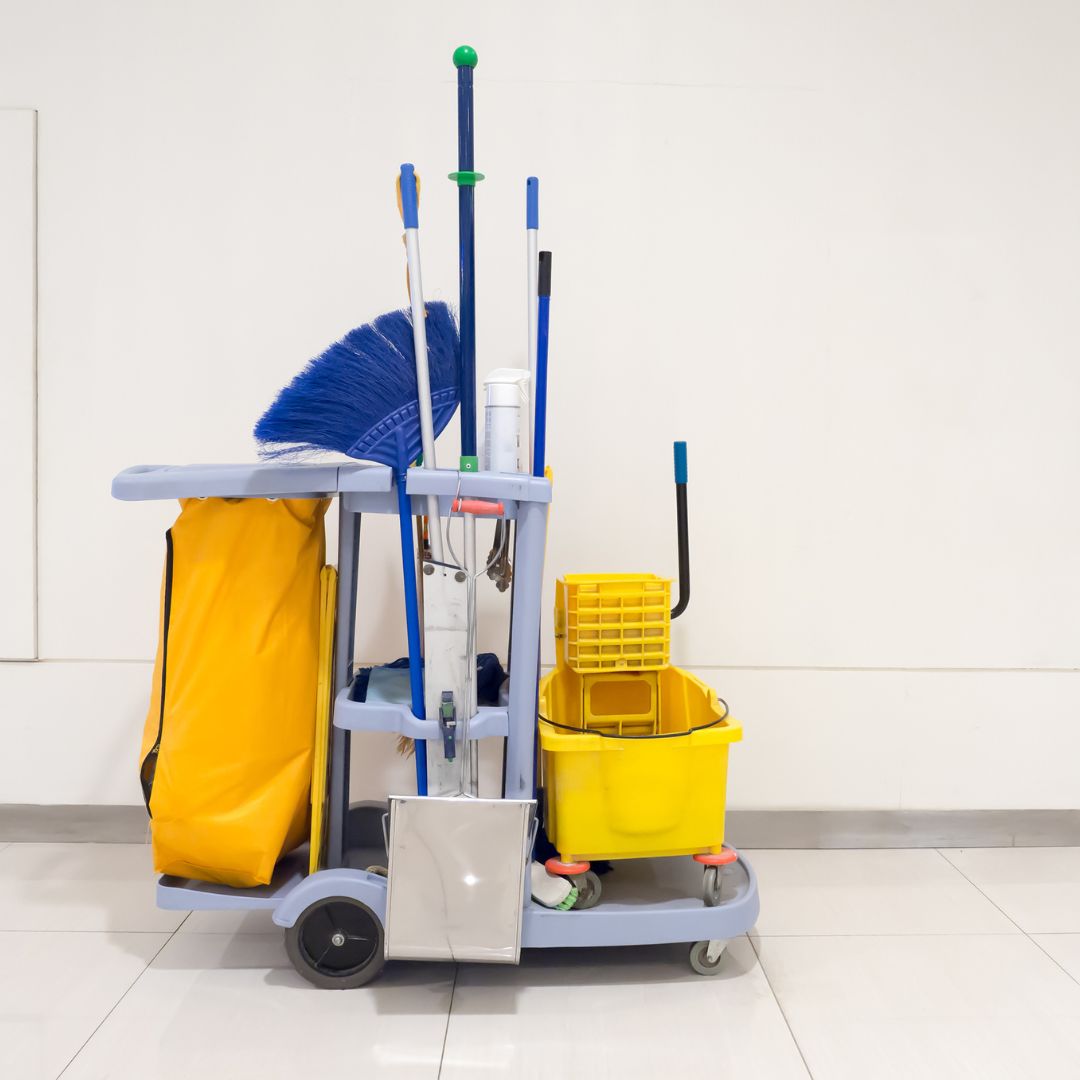 A professional cleaning cart with a yellow waste bag, mop bucket, brooms, and various cleaning tools is positioned against a plain white wall. The cart is organized and ready for use, with a tiled floor and simple background emphasizing the equipment.