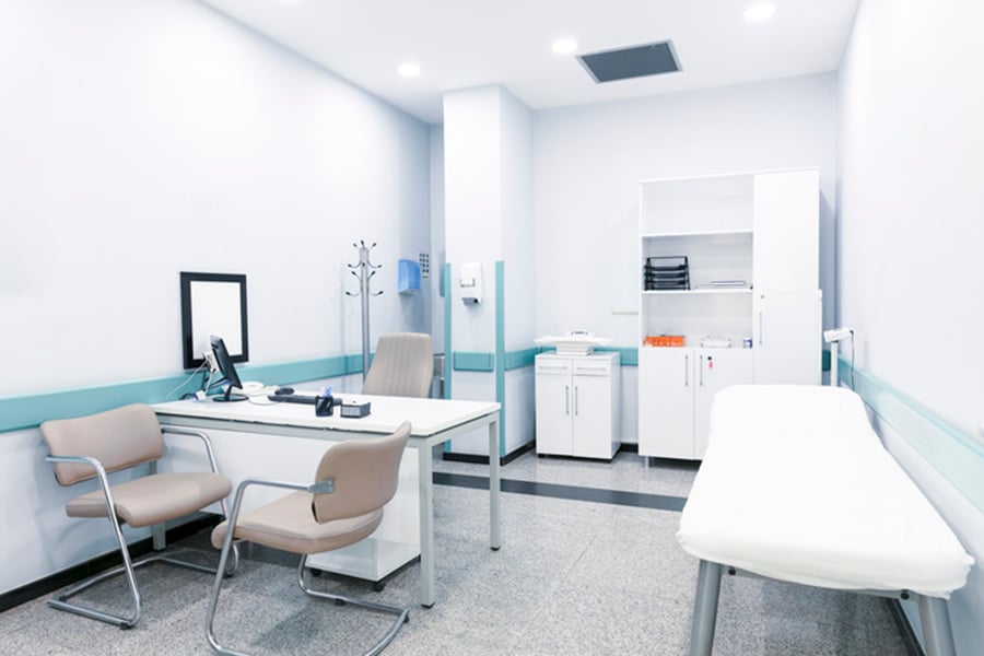 A medical examination room with a desk, two chairs, and an exam table, maintained by professional cleaning services.