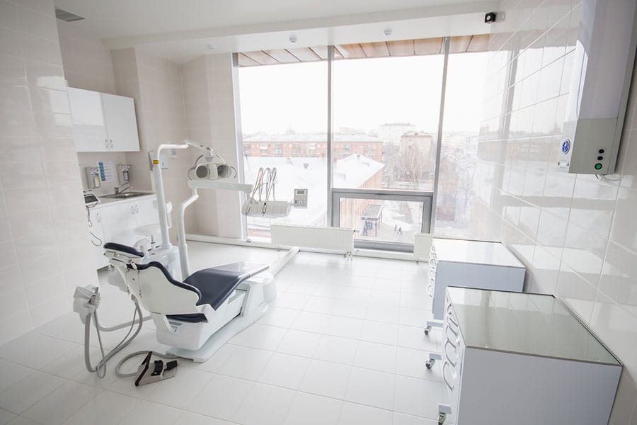 A bright, modern dental office that receives professional medical facility cleaning, featuring a reclining chair, dental equipment, and large windows overlooking an urban area.