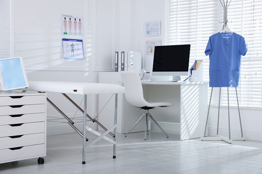 A bright medical examination room maintained through professional medical office cleaning, featuring a treatment table, a desk with a computer, and a blue scrub top hanging on a rack.