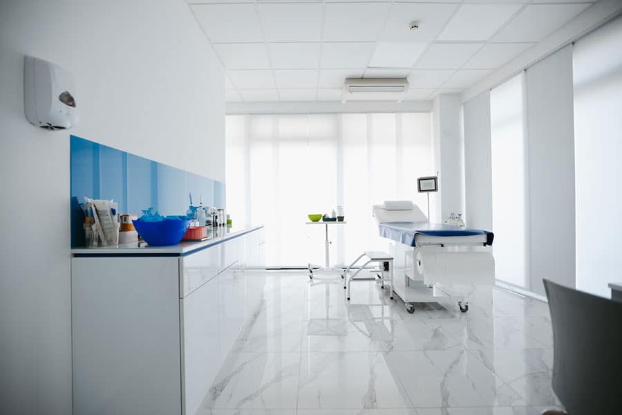 A sterile, brightly lit medical examination room maintained by professional medical cleaning, featuring a treatment table, a side counter with supplies, and white tiled floors.